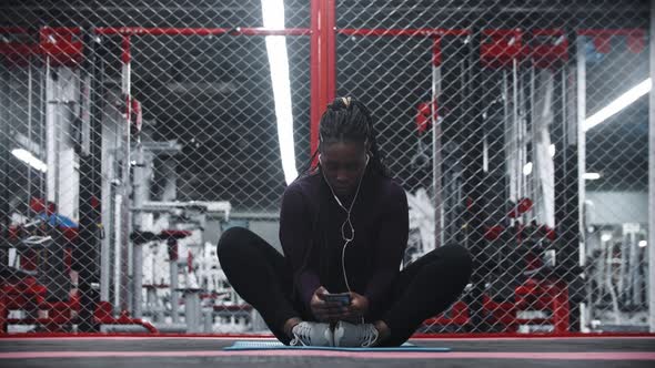 An Africanamerican Woman Sitting on Yoga Mat in the Gym  Using Her Phone and Listening Music