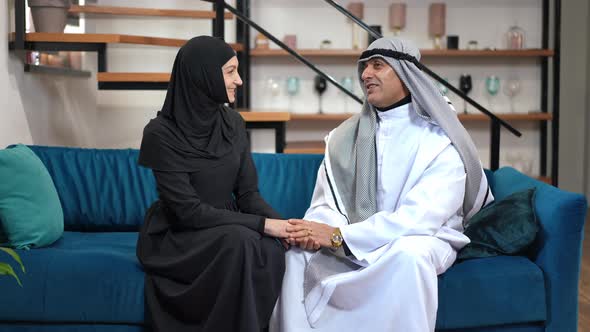 Happy Middle Eastern Couple Holding Hands Talking Sitting on Couch in Living Room