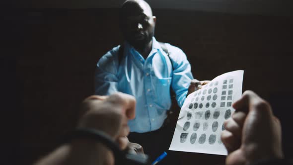 African Cop Showing Paper with Fingerprints To Suspect in Interrogation Room