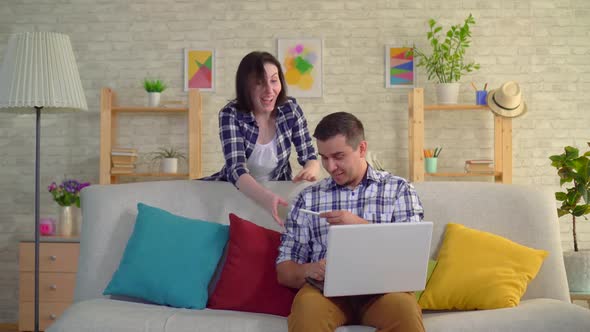 Young Positive Wife Dancing with Joy That She Is Pregnant Sad Husband in Shock Sitting at the Laptop