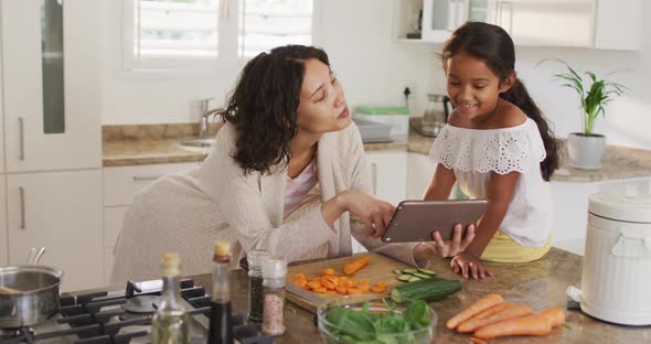 Hispanic mother teaching daughter sitting on countertop cooking, looking at tablet