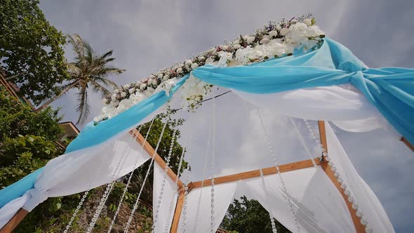 A Wedding Arch Decorated with Flowers and Large Winddeveloping Fabrics on a Tropical Beach