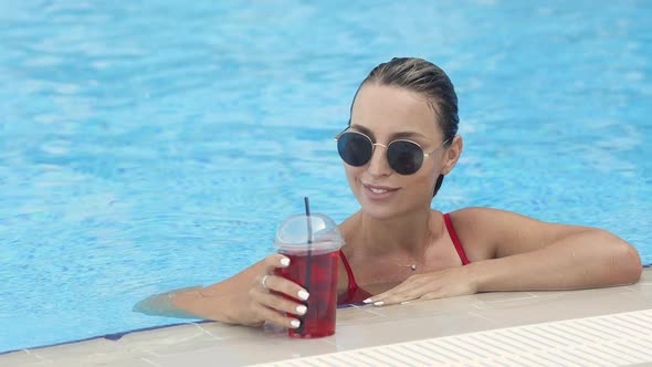 Beautiful smiling girl is resting by the pool in a red swimsuit with cold drink