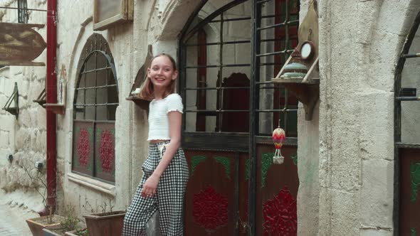 A Teenage Girl in Plaid Pants and a White Blouse Emerges From the Arch of the Doors of an Old