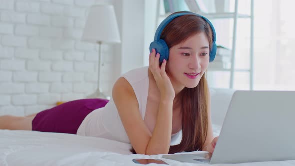 Happiness Asian young woman wear earphone lying on bed relax with music sing a song at home