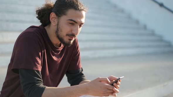 Young Attractive Arab Man Sitting in Park and Using Smartphone