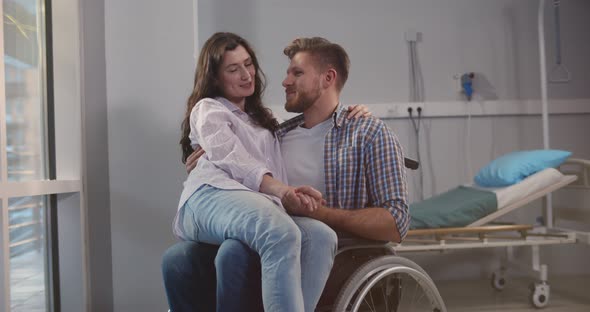 Smiling Woman Sitting on Lap of Disabled Boyfriend in Wheelchair at Hospital