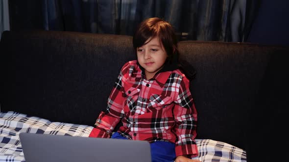 Boy Child in a Red Plaid is Watching a Computer Sitting on Bed at Home