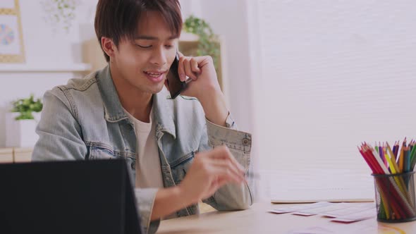 In his living room, an Asian man is on the phone, listening and conversing on his smartphone.