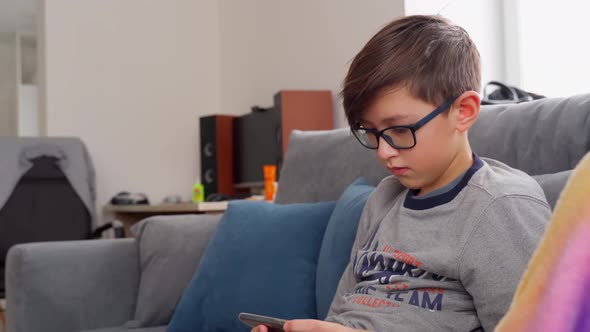 A Boy with Glasses Sits on the Couch and Plays on the Phone