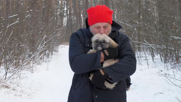 Aged Man in Red Hat Holds Dog in Arms and Walks Along Forest