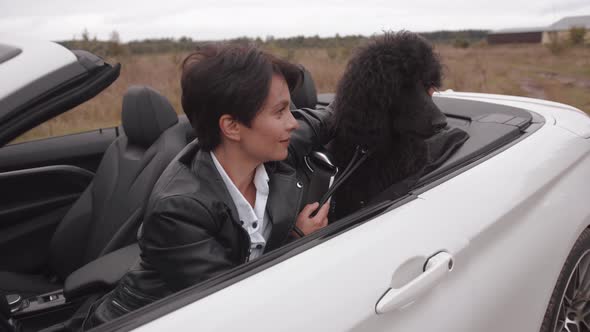 Stylish Young Woman in a Black Leather Jacket Sits in a Convertible and Strokes a Large Poodle