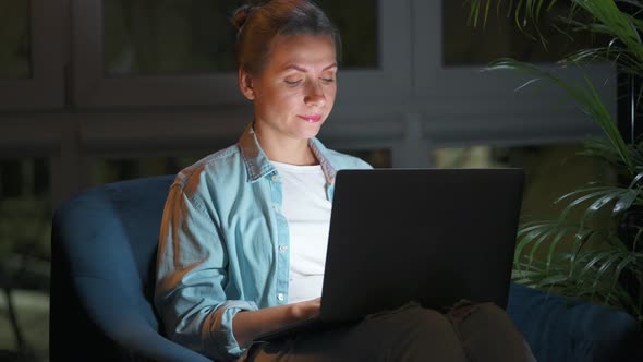 Woman is Sitting in the Armchair and Working on a Laptop at Night