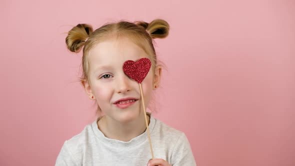 Funny Little Blonde Girl Smiling and Playing with Red Heart Toy on Pink Background