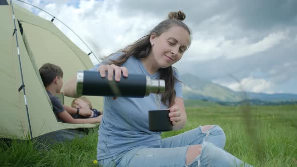 Smiling millennial woman in blue t-shirt pouring hot tea from thermos against tent with kids