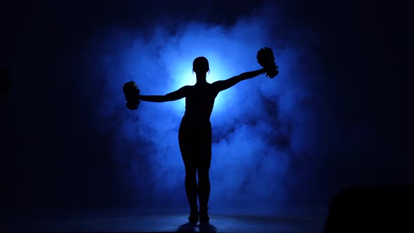 Dark Silhouette of Cheerleader with Pompoms in Her Hands Against Background of Smoke and Spotlight