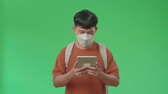 Asian Boy Student Wearing A Mask, Using Tablet While Walking On Green Screen