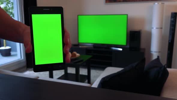 A Hand Holds a Smartphone with a Green Screen - Closeup, TV with a Green Screen in the Background