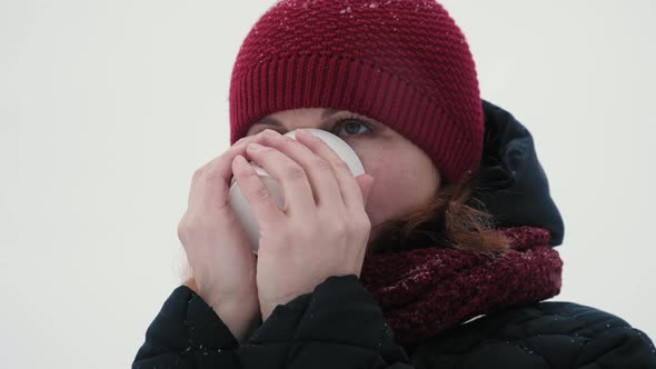 Woman in a red warm hat is drinking hot tea or coffee from a cup in a snowy forest