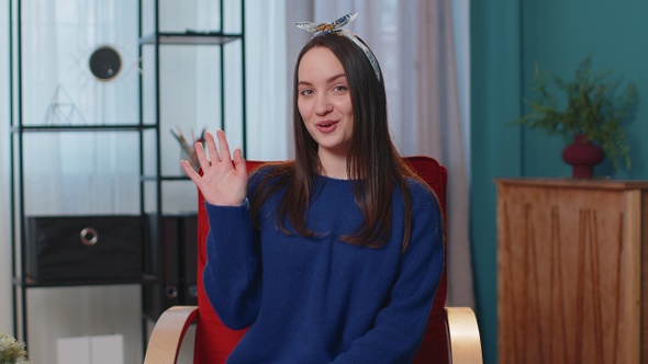 Young Woman Smiling Friendly at Camera Waving Hands Gesturing Hello Hi Greeting at Home on Chair