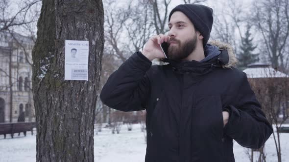 Young Bearded Caucasian Man Talking on the Phone and Looking at Missing Person Ad Hanging on the