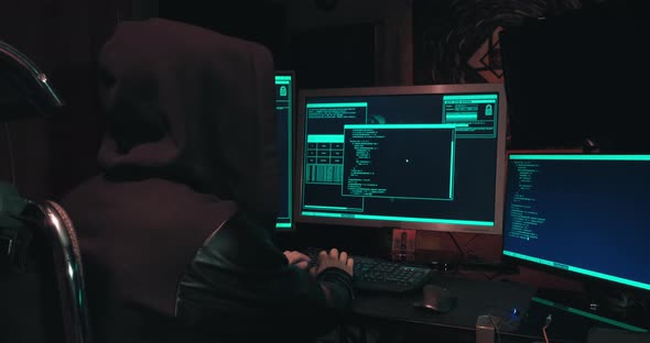 Hacker in a Hood Sits in Front of Computer Screens and Hacks Databases