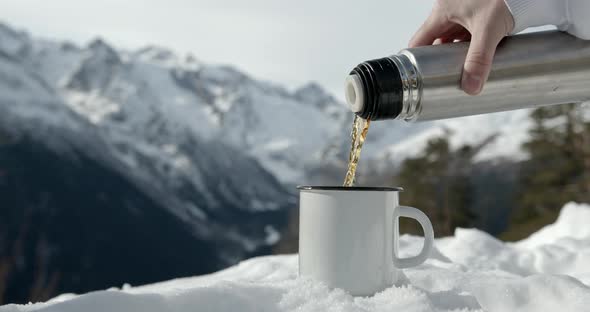 Pouring hot liquid coffee from a thermo into a cup during a cold winter with mountains full of snow