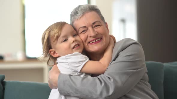 A Little Blondie Boy Kisses and Hugs a Mature Woman in a Gray Jacket Sitting on the Couch