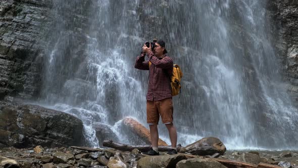Travel Man with a Yellow Backpack Standing on the Background of a Waterfall Makes a Photo Landscape