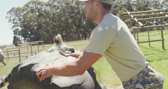 Fit caucasian male soldier in cap rolling tractor tyre on obstacle course in the sun