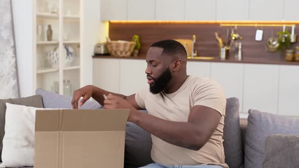 Excited African American Happy Guy Received a Parcel at Home Unpacks a Cardboard Box Examines the