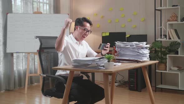 Happy Asian Man Celebrating Winning Game On Smartphone After Working With Documents At The Office