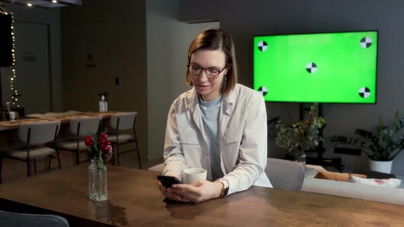 A beautiful girl with glasses sits at a table in the living room In the background is a green screen