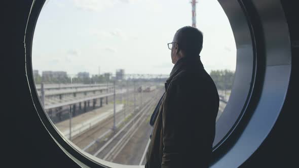 Elegant Mature Man Standing at Railway Station and Looking Out of Round Window at Tracks