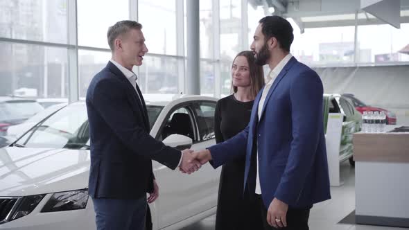 Adult Car Dealer Welcoming Young Multiracial Couple in Showroom. Successful Middle Eastern Man and