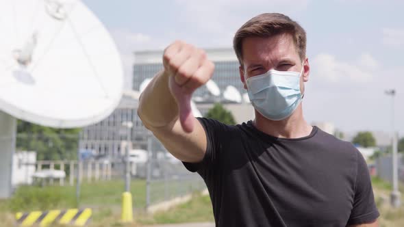 A Young Man in a Face Mask Shows a Thumb Down To the Camera and Shakes His Head in an Urban Area