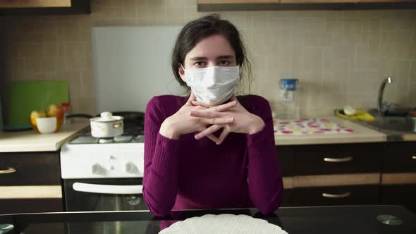 Girl removes a medical mask while sitting at home, in the background of the kitchen