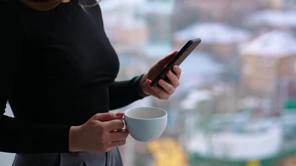 Beautiful female with cup of coffee thoughtfully browsing smartphone while standing by the window