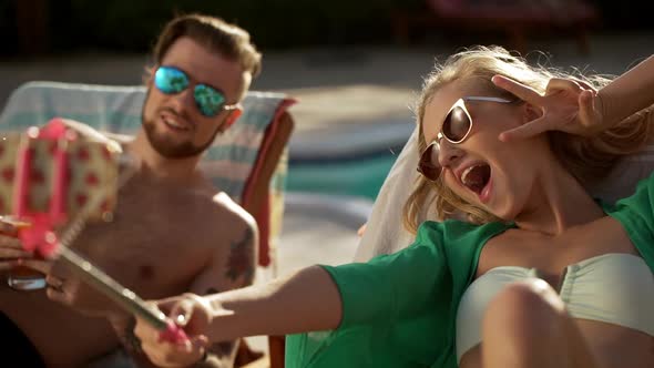 Attractive Blonde Female in Sunglasses Making Selfies with Mobile Phone and Stick While Topless Male