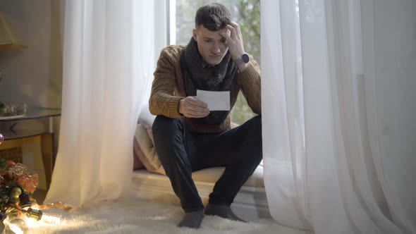 Wide Shot of Depressed Young Man Sitting on Windowsill Looking at Ultrasound Picture