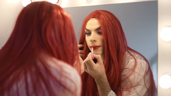 Confident Transgender Woman in Red Wig Applying Red Lipstick Reflecting in Mirror