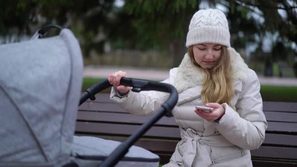 Happy Mother with Smartphone Using App. She Sitting on the Bench , Baby Sleeping in Pram