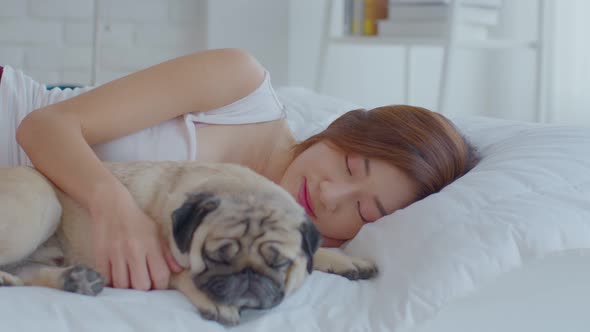 Beautiful Asian young woman hug cute dog pug breed and sleep together in cozy white bedroom