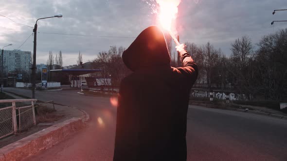 Young Man in Hoodie and Balaclava with Red Burning Signal Flare on the Road Under an Old Steel Frame