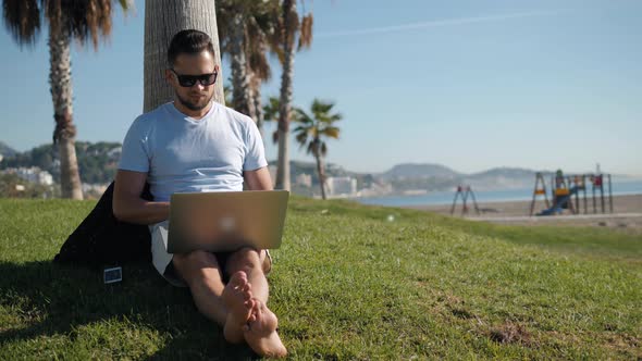 Handsome Tourist Man Student in Glasses Hipster Using Laptop in Park on a Summers Day Sitting on