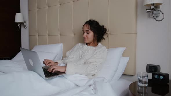 The Woman Works on a Laptop Computer and Sits on the Bed