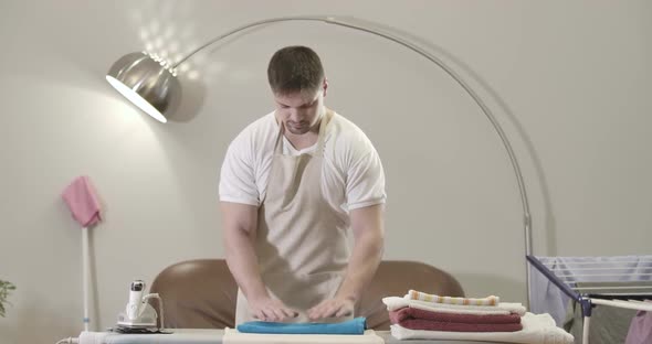 Portrait of Happy Young Caucasian Man Folding Clean Laundry on Ironing Board. Cheerful Handsome Guy