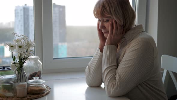 Unhappy Woman Sitting At Table In Kitchen And Holding Head From Migraine Pain