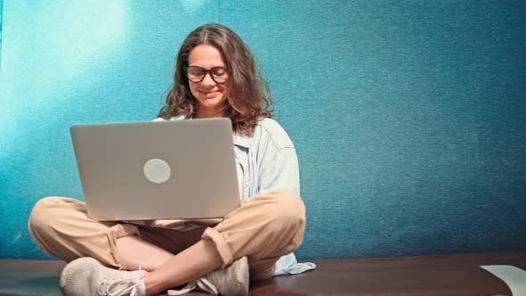 A Young Cheerful Woman in Glasses is Typing on a Laptop