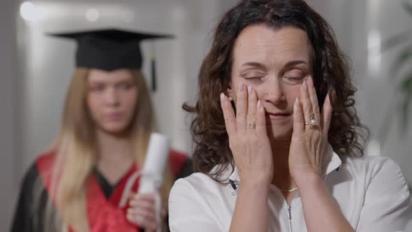 Mature Caucasian Woman Crying As Blurred Graduate Daughter in Gown and Hat Standing at Background